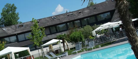 Una piscina con sillas de descanso y sombrillas, rodeada por un edificio con amplias ventanas y una palmera.