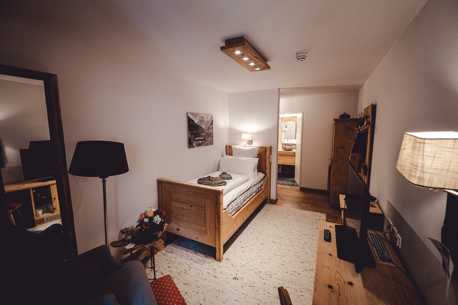 A cozy bedroom with a wooden bed, a desk, a chair, a lamp, and a mirror.