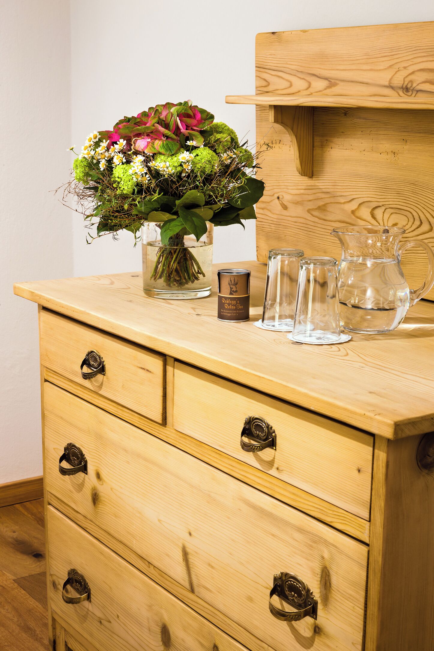 A wooden dresser with a vase of flowers, a candle, and two glasses on top.