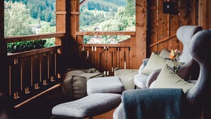 A cozy wooden cabin with a view of a forest and a distant building. There are two upholstered chairs with cushions and a small table with a vase of flowers. - Berghotel Rehlegg (Ramsau bei Berchtesgaden)