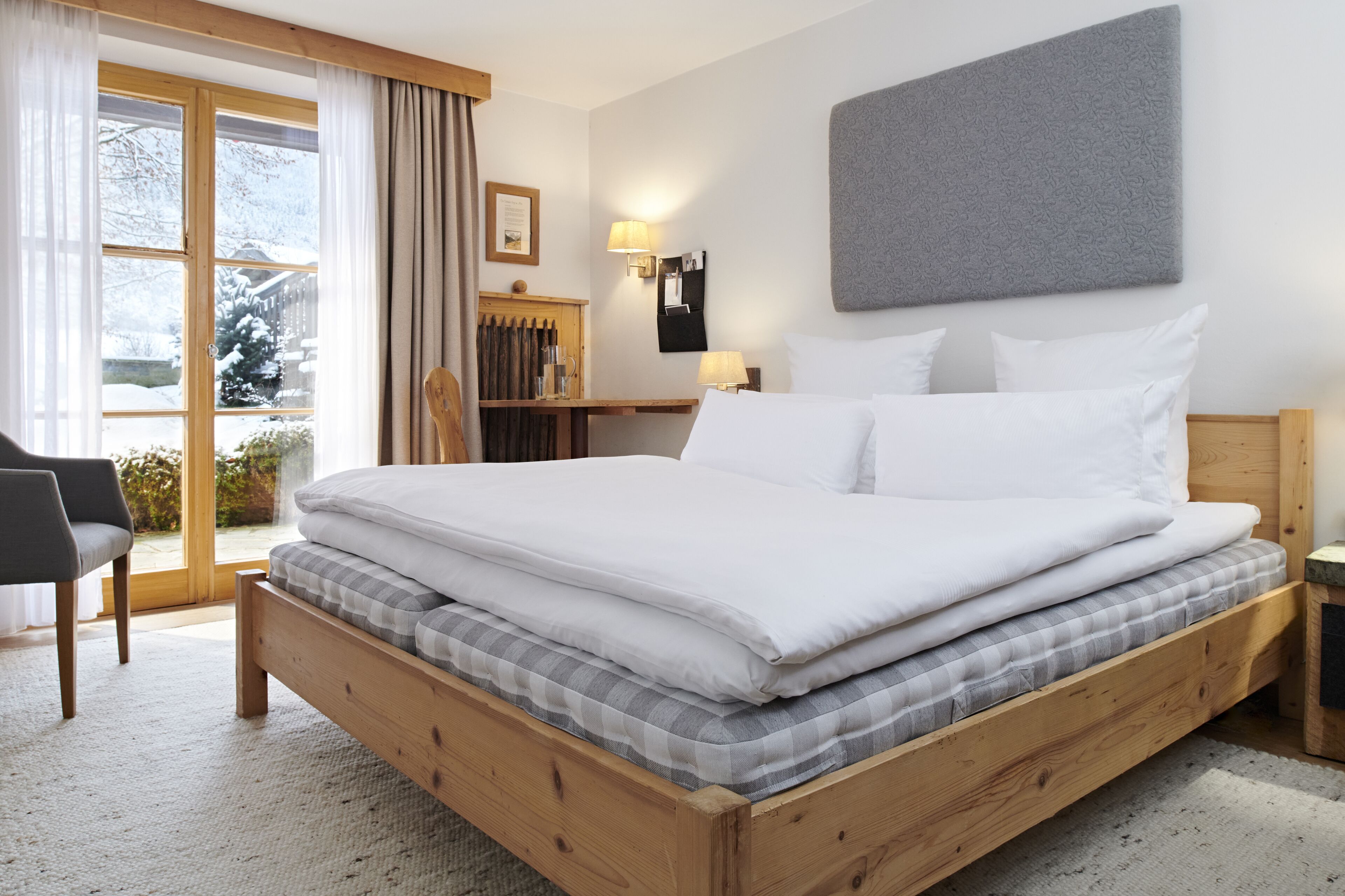 A wooden bed with white and gray bedding, a wooden nightstand, a chair, a window with curtains, and a wall-mounted lamp.