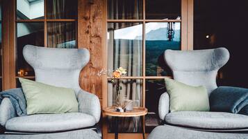 Zwei Sessel mit grünen Kissen, ein kleiner Holztisch mit Blumenvase und Blick auf eine Berglandschaft durch große Fenster.