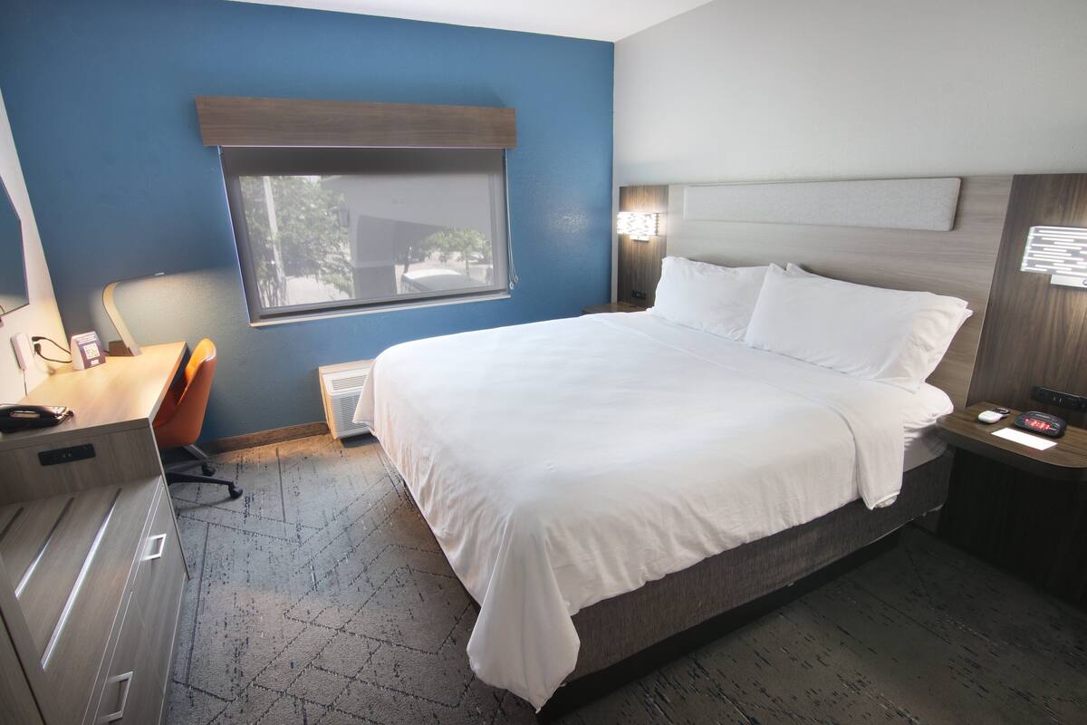A hotel room with a large bed, a desk with a chair, a window, and a nightstand with a lamp.