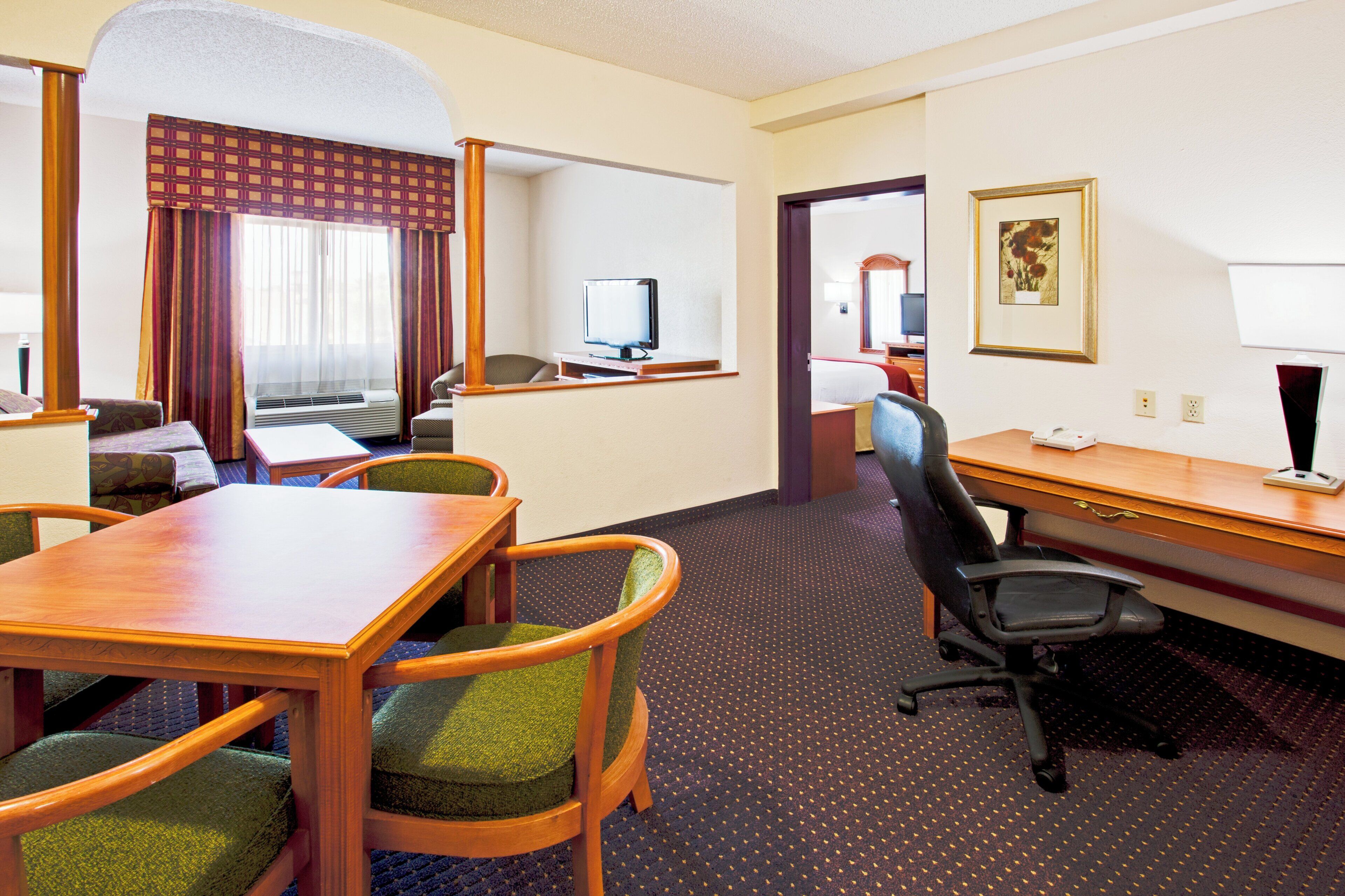 A hotel room with a dining table, chairs, a desk, a computer, and a television.