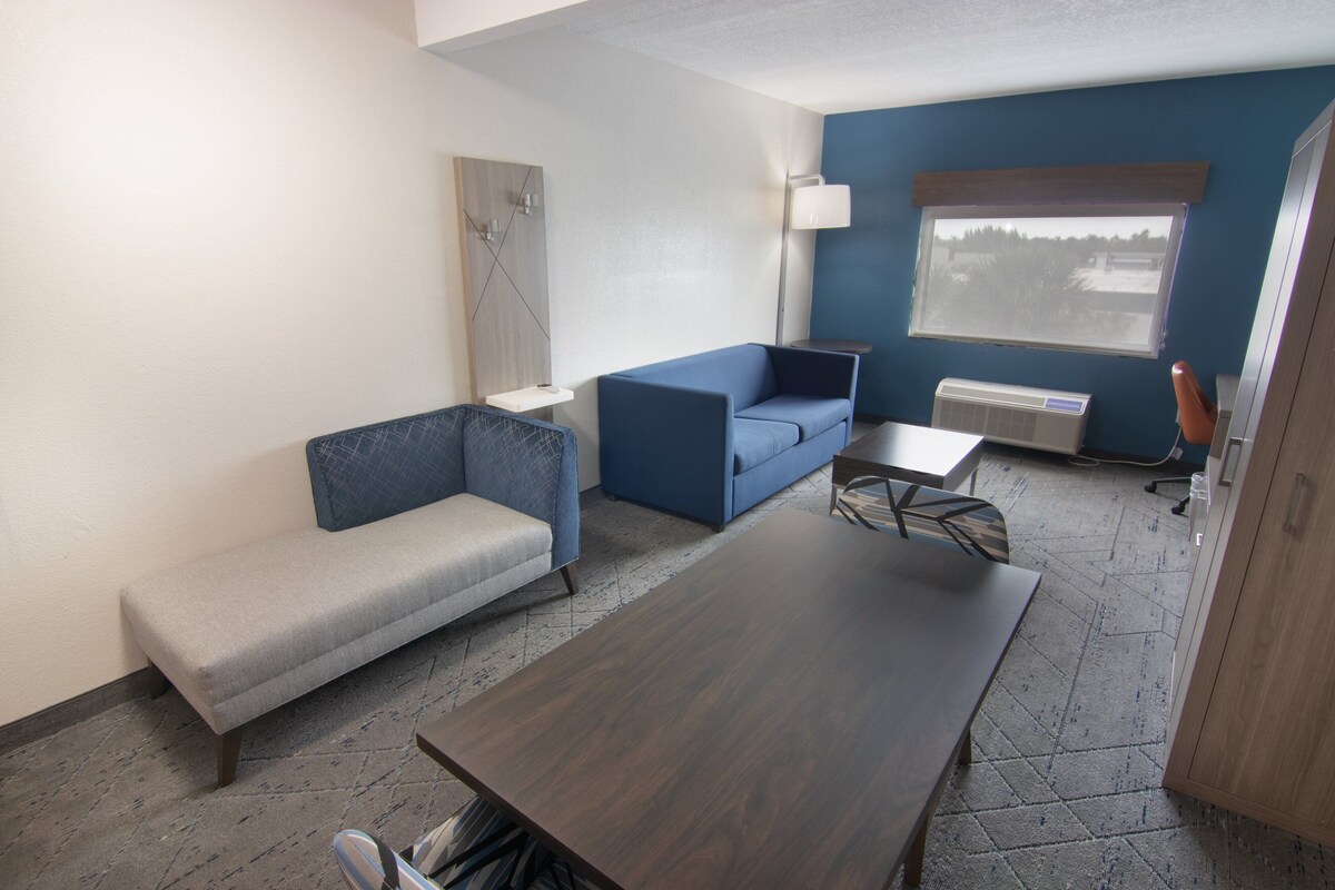 Una habitación moderna con sofá gris, sillón azul, mesa de madera y ventana con vistas a árboles.