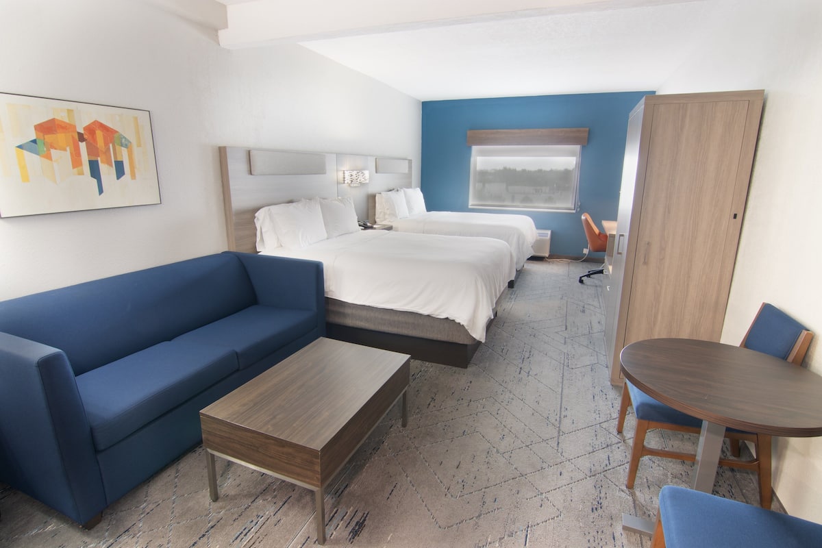 A hotel room with a blue sofa, a wooden coffee table, a bed with white bedding, a desk with a chair, and a window with a view.