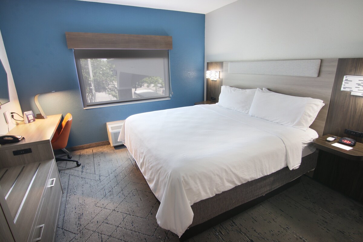 Habitación de hotel con una cama grande, un escritorio con silla, una ventana y una mesita de noche con lámpara.