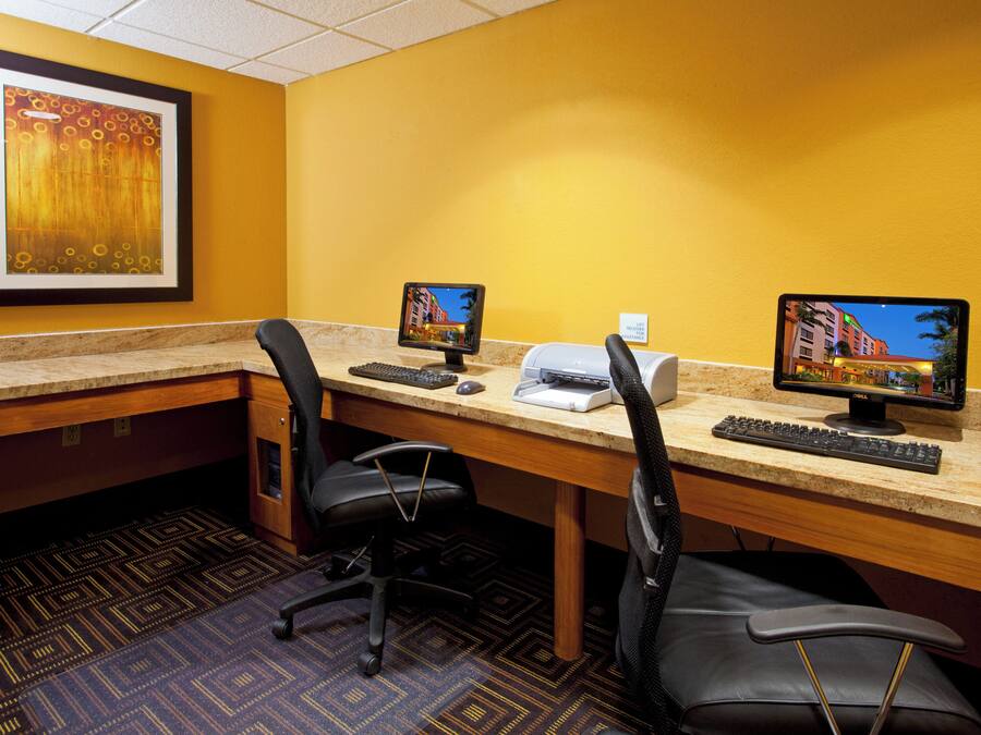 A hotel room with two desks, each with a computer, keyboard, and mouse. There is a framed picture on the wall and a carpeted floor.