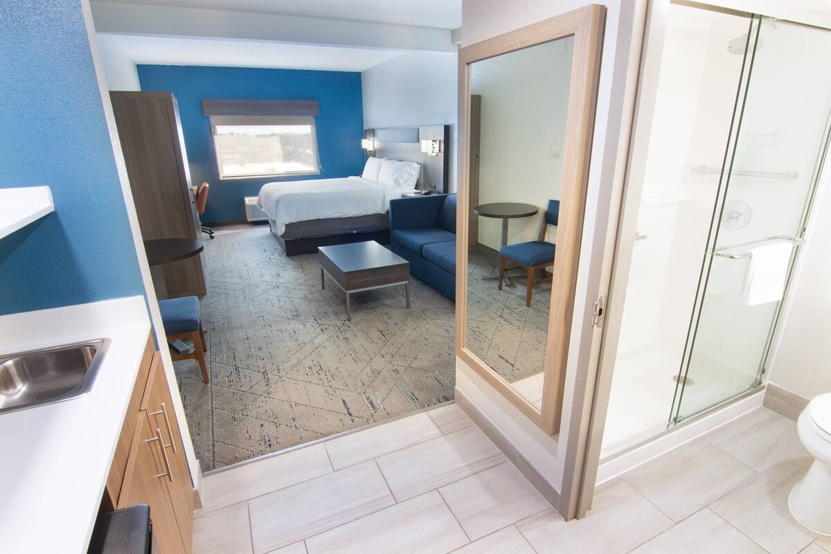A hotel room with a bed, a sofa, a small table, a mirror, a bathroom with a shower, and a toilet.