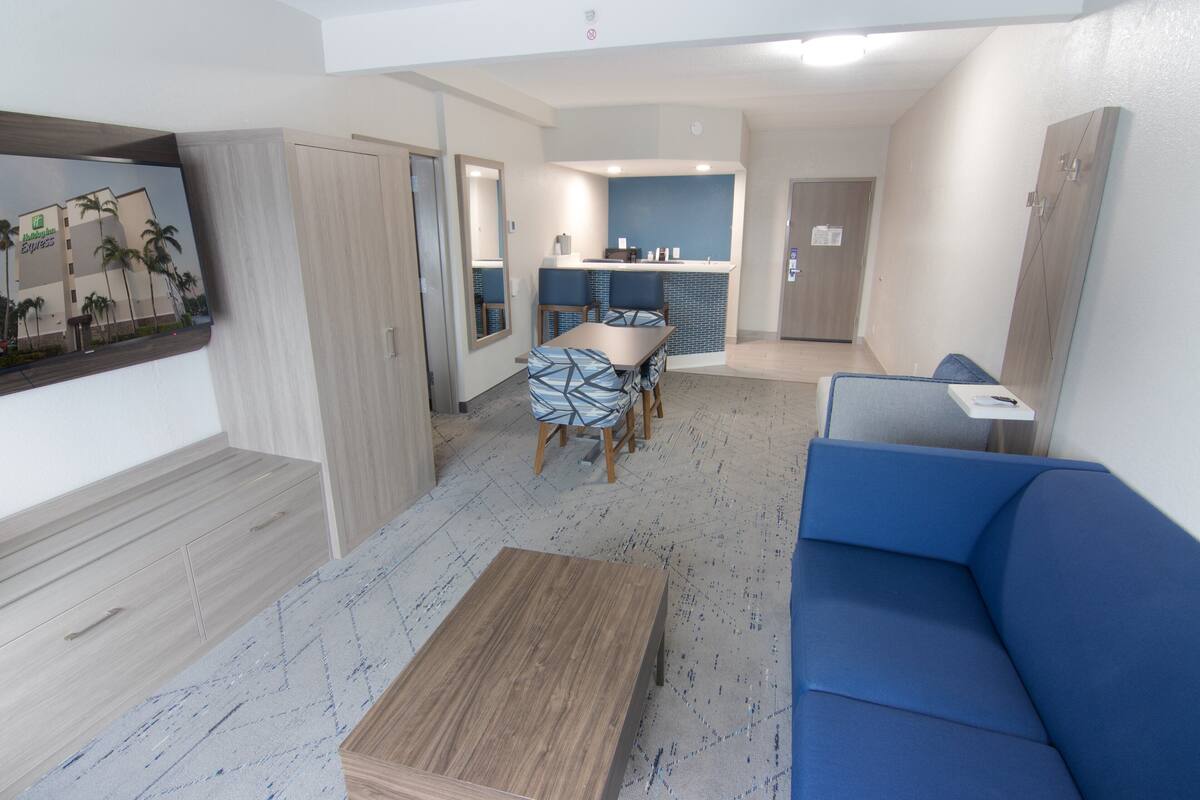 A modern living room with a blue sofa, wooden coffee table, and patterned chairs. A kitchen area is visible in the background with a blue backsplash and wooden cabinets.