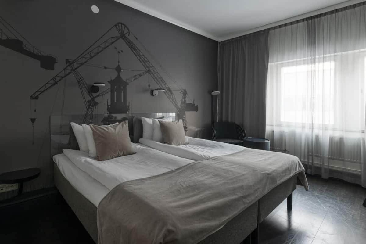 Habitación de hotel con dos camas, un ventanal con cortinas translúcidas, un mural de una grúa y una mesita de noche.