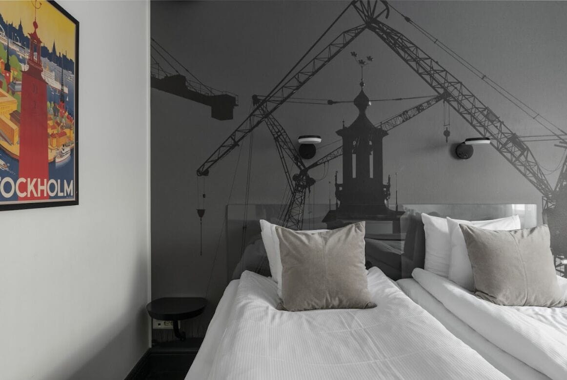 A hotel room with a bed, pillows, a nightstand, a wall-mounted lamp, and a poster of a cityscape.