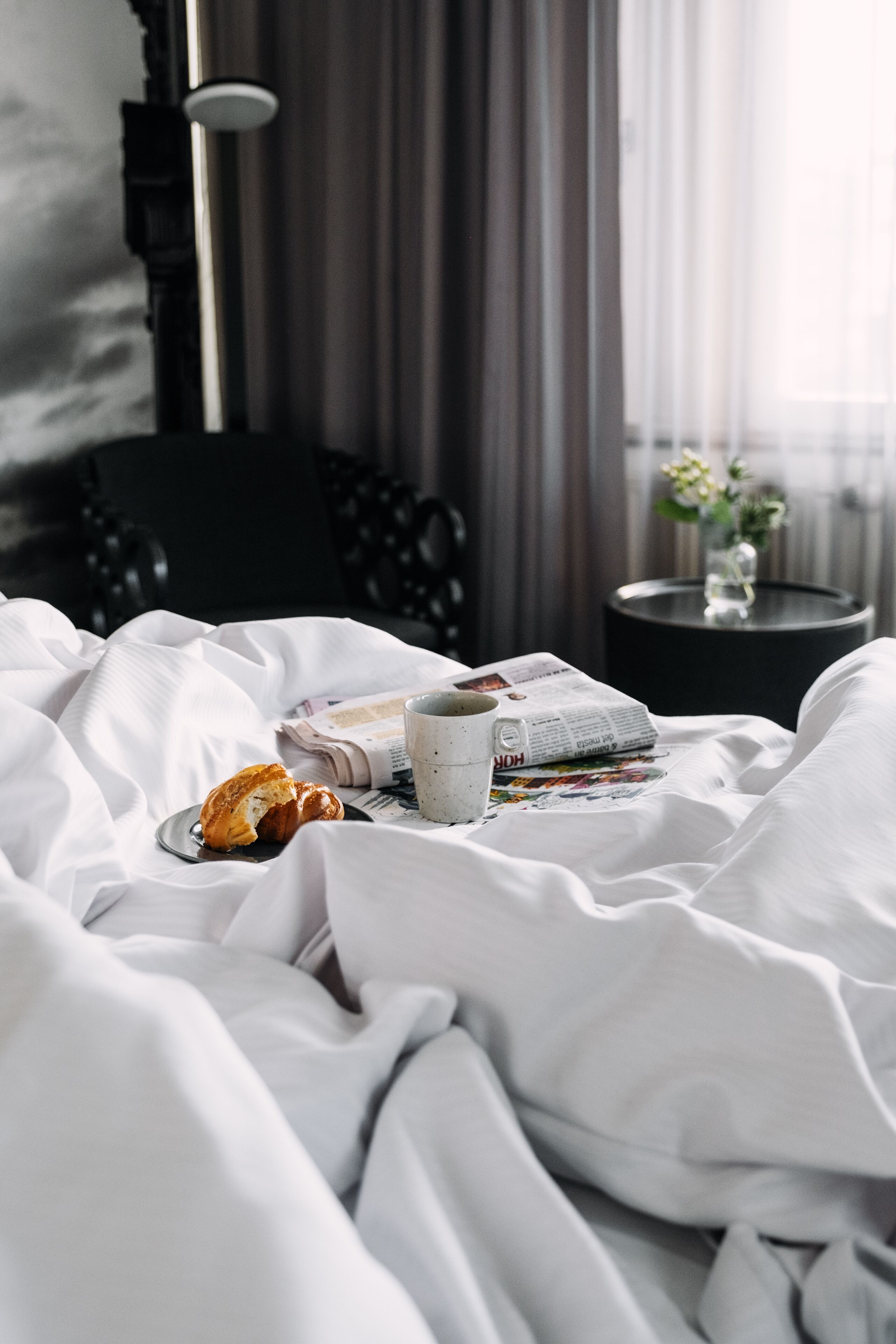 Ein ordentlich bezogenes Bett mit einem Croissant, einer Tasse und einer Zeitung. Im Hintergrund steht ein kleiner Tisch mit einer Blumenvase.