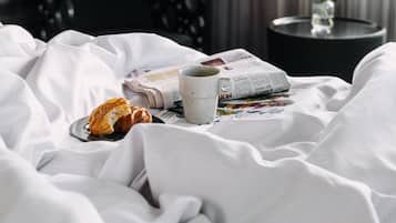 Ein ordentlich bezogenes Bett mit einem Croissant, einer Tasse und einer Zeitung. Im Hintergrund steht ein kleiner Tisch mit einer Blumenvase.