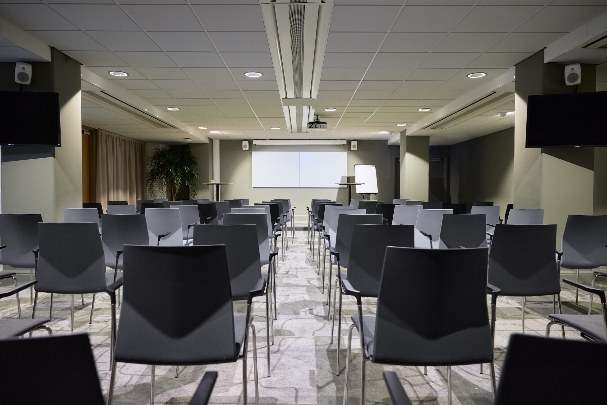 Une salle de conférence avec des rangées de chaises disposées face à un écran de projection, un tableau blanc et un haut-parleur mural.