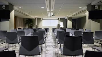 A conference room with rows of chairs facing a projection screen, a whiteboard, and a speaker on the wall.