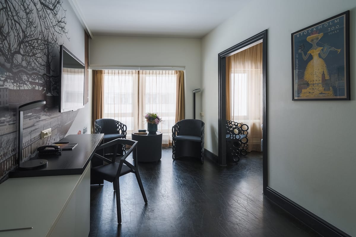 Una habitación de hotel moderna con piso de madera oscura, un escritorio con teléfono, una silla, una mesita con un florero y un ventanal con cortinas translúcidas.