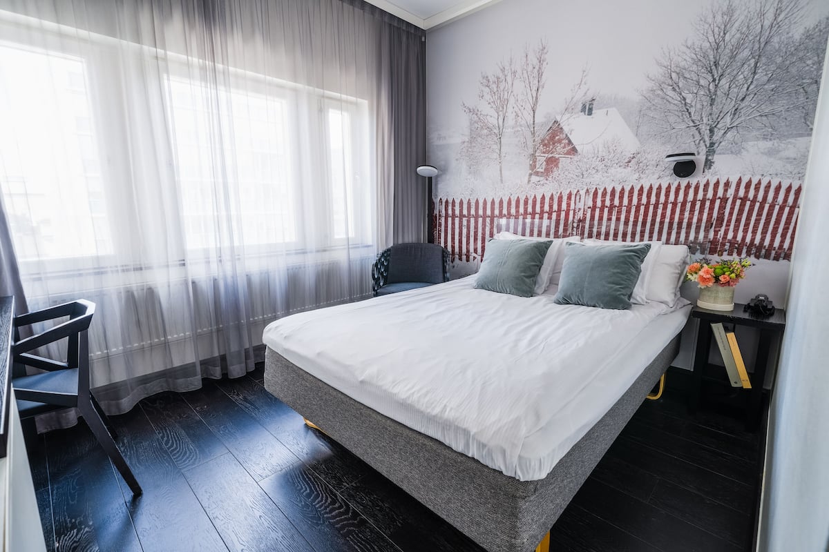 Un dormitorio con una cama, una silla, una mesita de noche, una lámpara y una ventana con cortinas translúcidas.