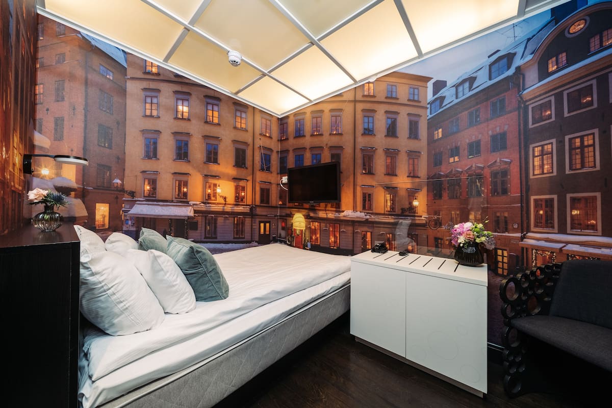 Habitación de hotel moderna con una cama, un escritorio y un televisor grande. Se aprecia una vista panorámica de la ciudad a través de las ventanas.
