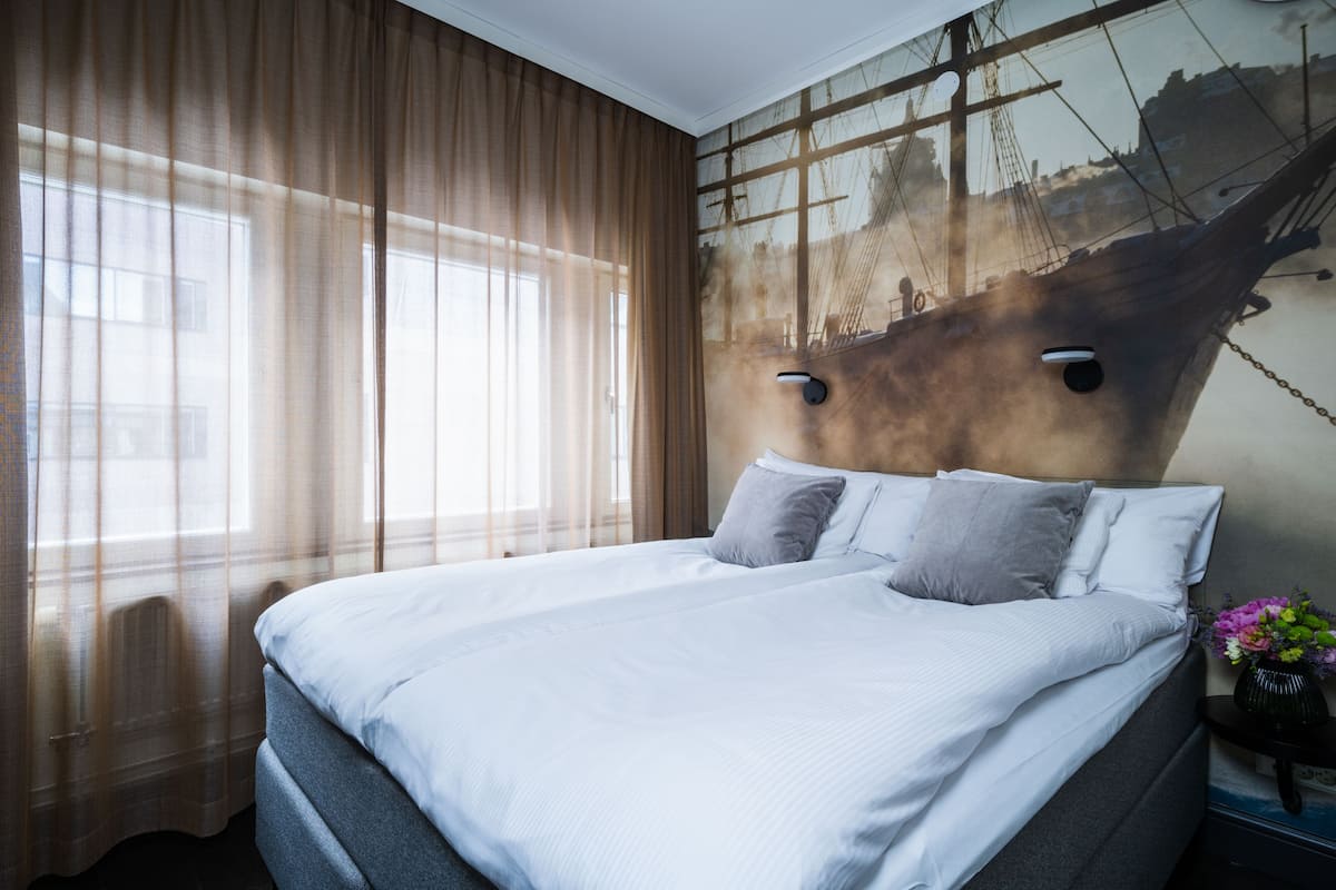 Una habitación de hotel moderna con una cama grande, cortinas translúcidas, un mural temático de barcos y una mesita de noche con un florero.