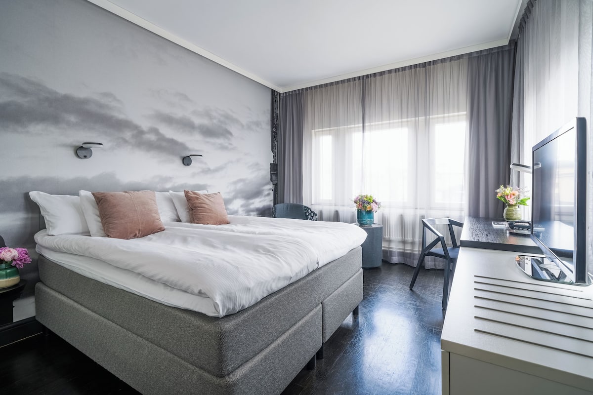 Una habitación de hotel moderna con una cama grande, un escritorio con computadora y un ventanal con cortinas.