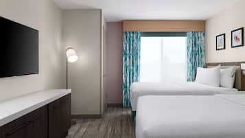 Quarto de hotel com duas camas, televisão de tela plana, escrivaninha com gavetas, luminária e janela com cortinas.