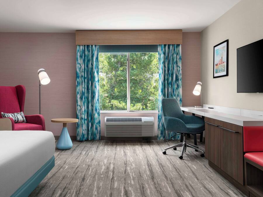 Habitación de hotel con cama, escritorio, silla, televisión y ventana con cortinas.