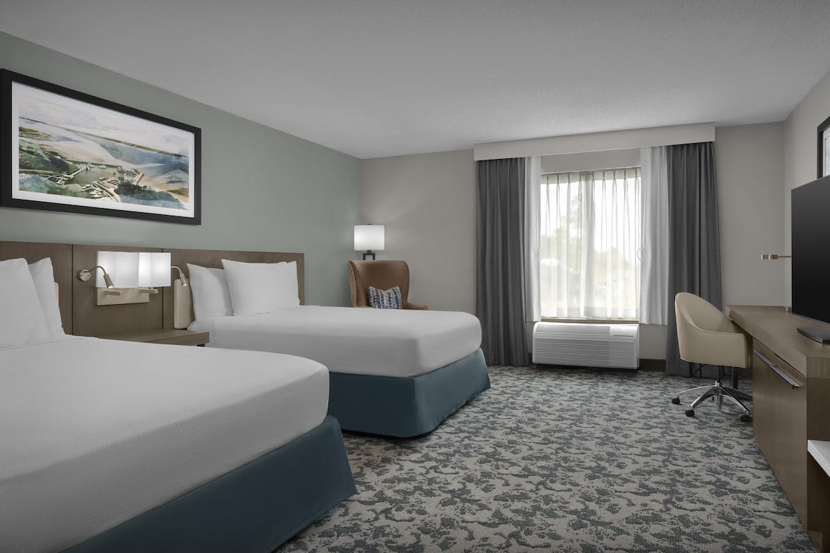 A hotel room with two beds, a desk, a chair, a TV, a lamp, a painting, and a window with curtains.