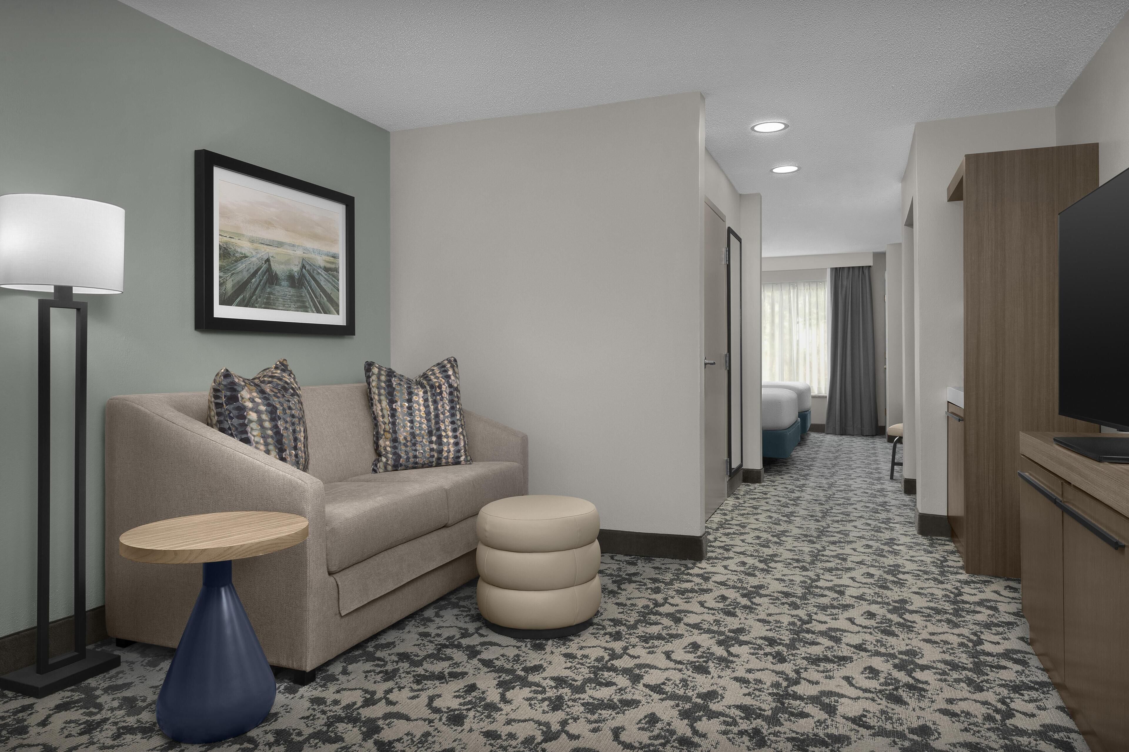 A hotel room with a sofa, ottoman, lamp, framed picture, and a television.