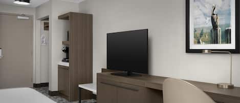A hotel room with a bed, a desk with a TV, a chair, a framed picture on the wall, and a door.