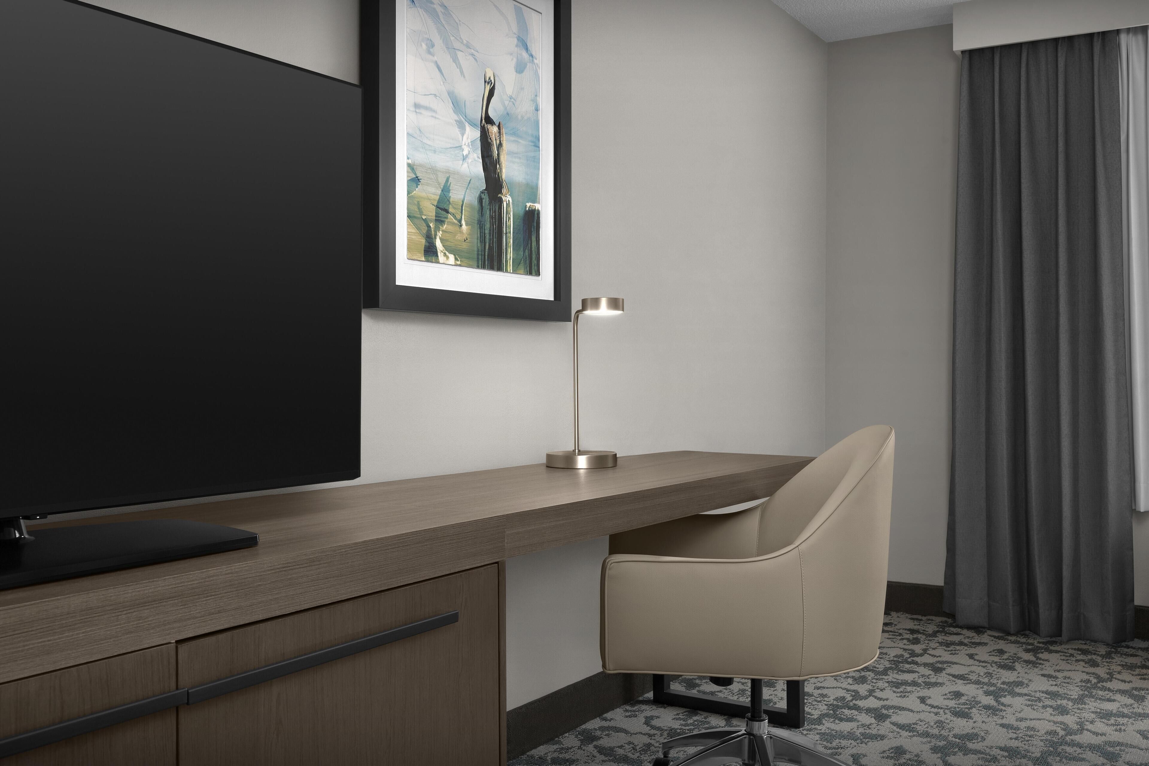 A hotel room with a flat-screen TV, a desk with a lamp, a chair, and a framed picture on the wall.