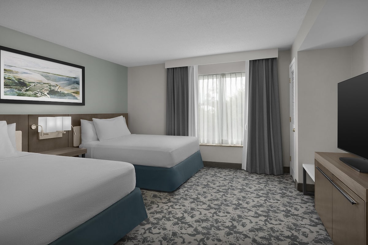 A hotel room with two beds, a television, a painting, and a window with curtains.
