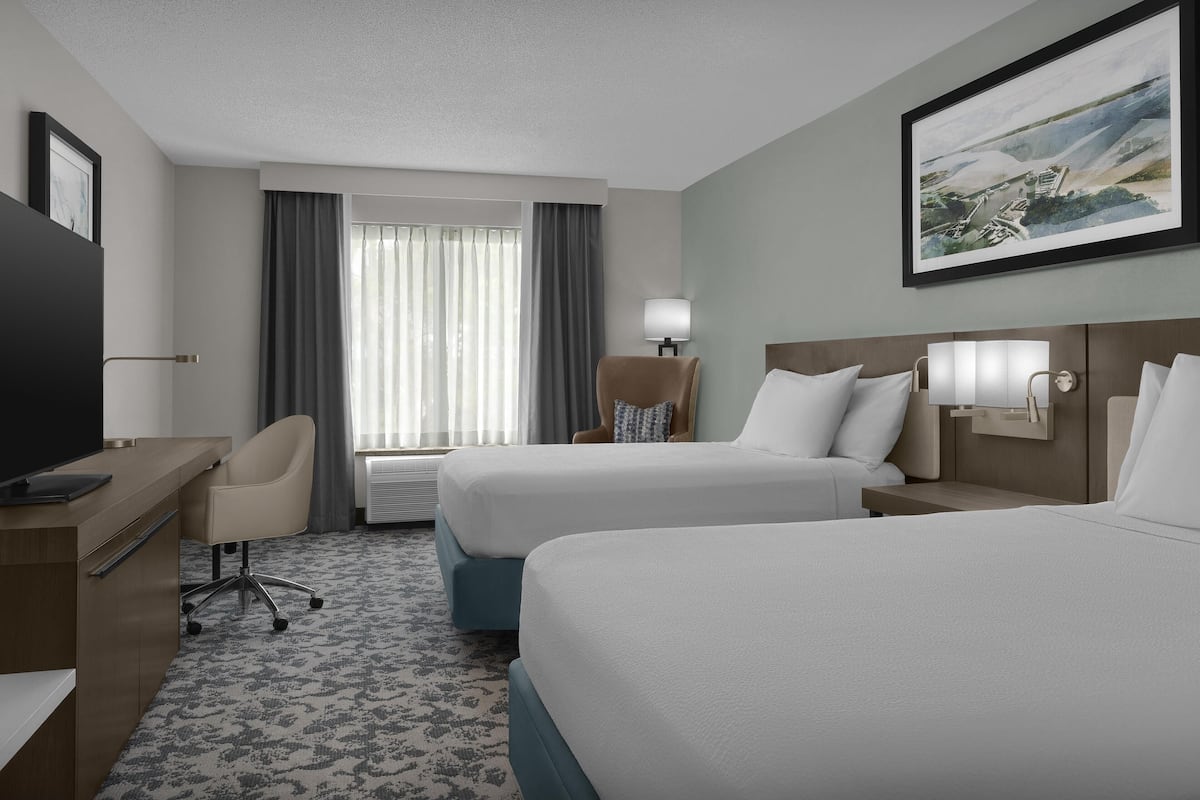 A hotel room with two beds, a desk, a chair, a TV, and a painting on the wall.