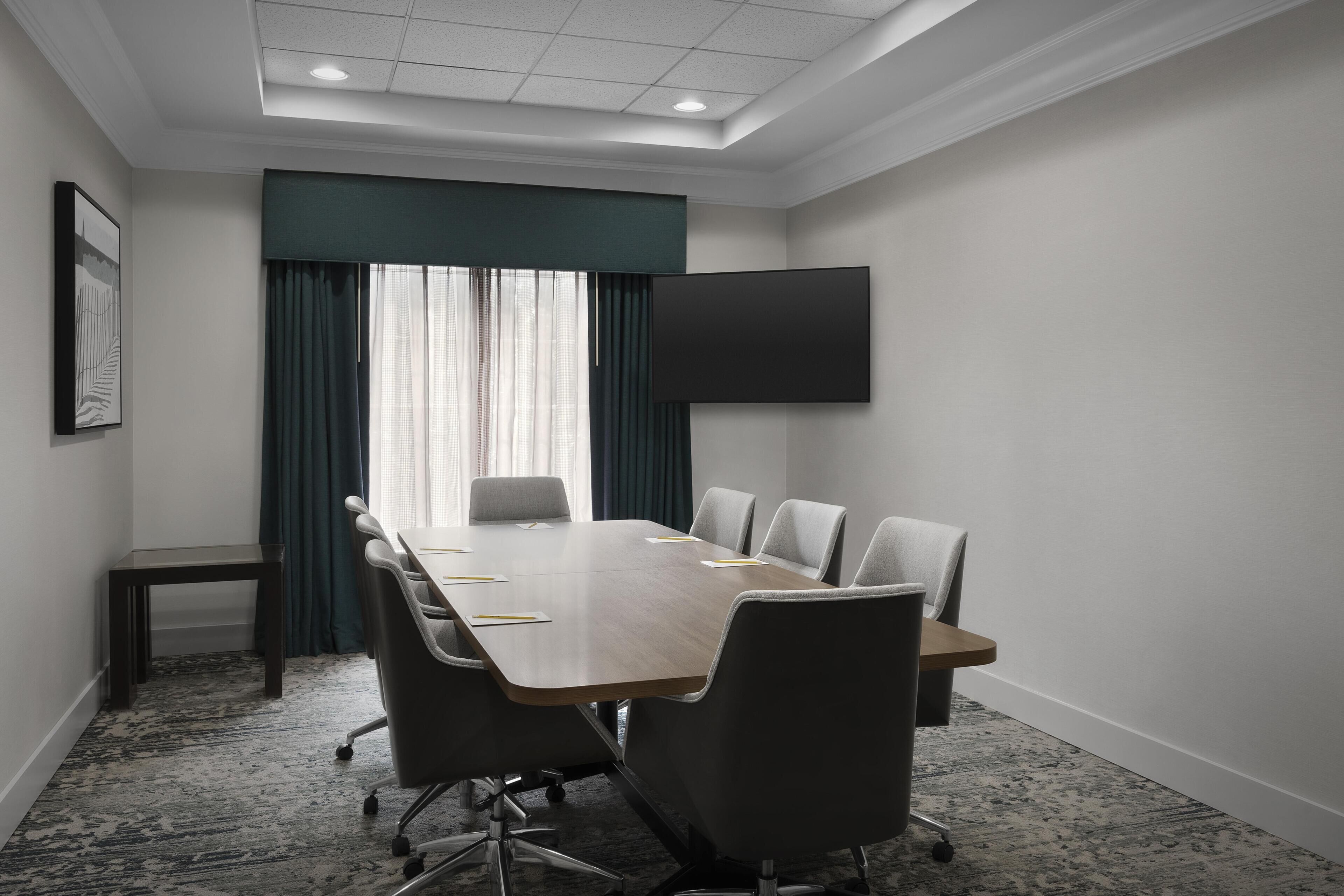 A conference room with a long table, six chairs, a TV mounted on the wall, and a window with curtains.