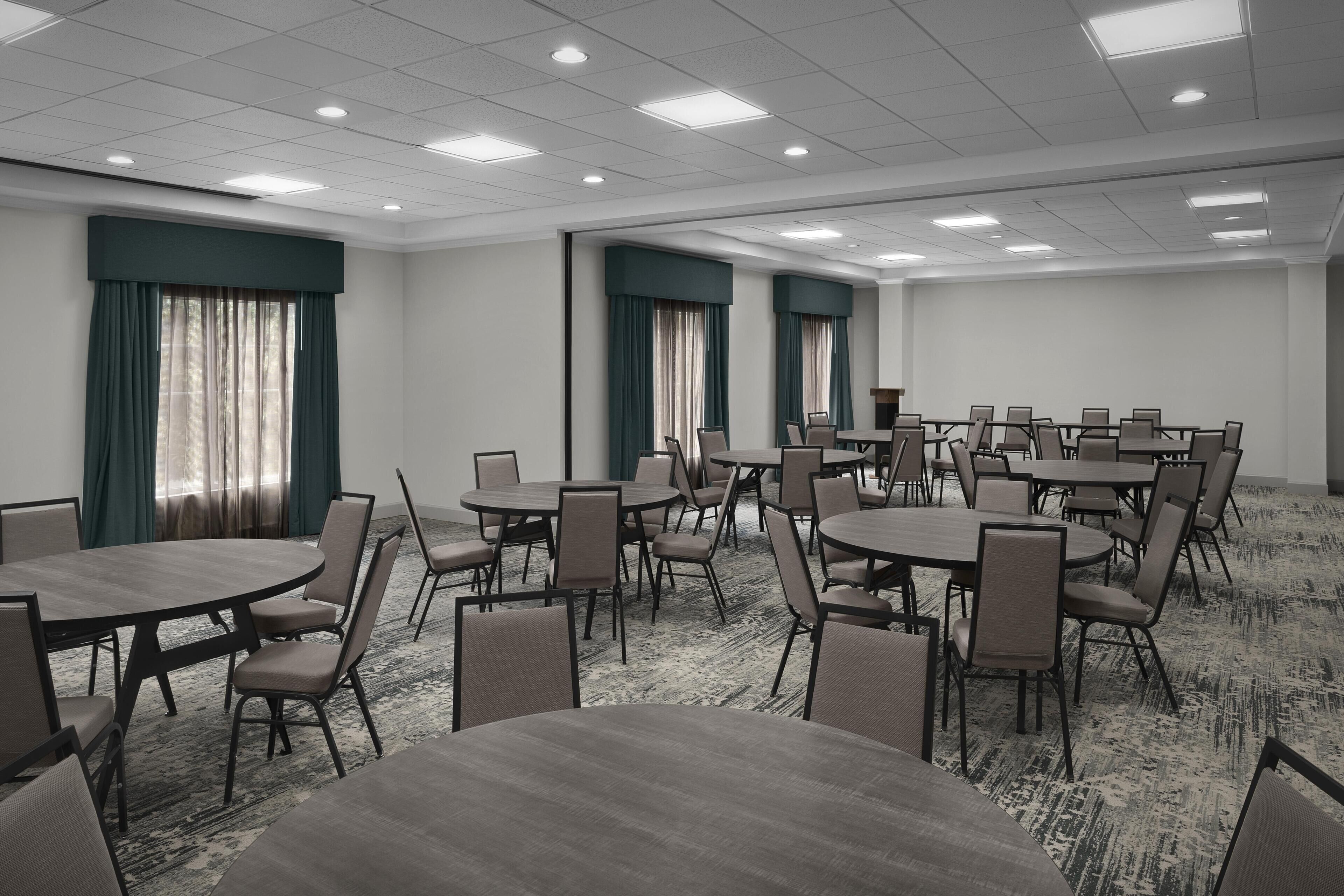 A conference room with multiple tables and chairs, large windows with curtains, and a carpeted floor.