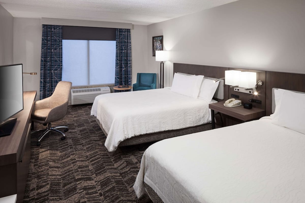 A hotel room with two beds, a desk with a television, a chair, a lamp, and a bedside table with a phone and alarm clock.