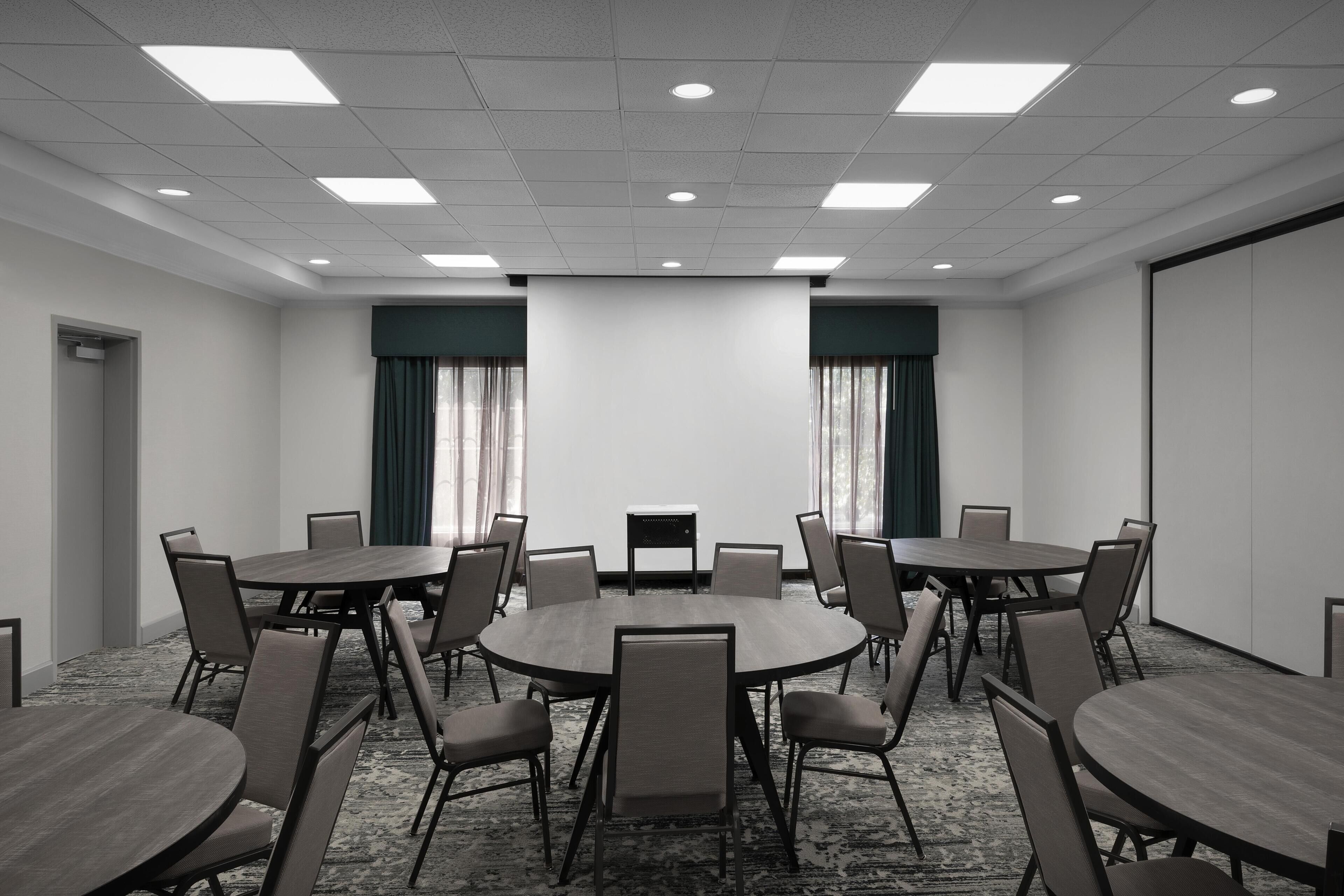Une salle de conférence équipée de tables rondes et de chaises, d’un écran de projection et d’un sol recouvert de moquette.