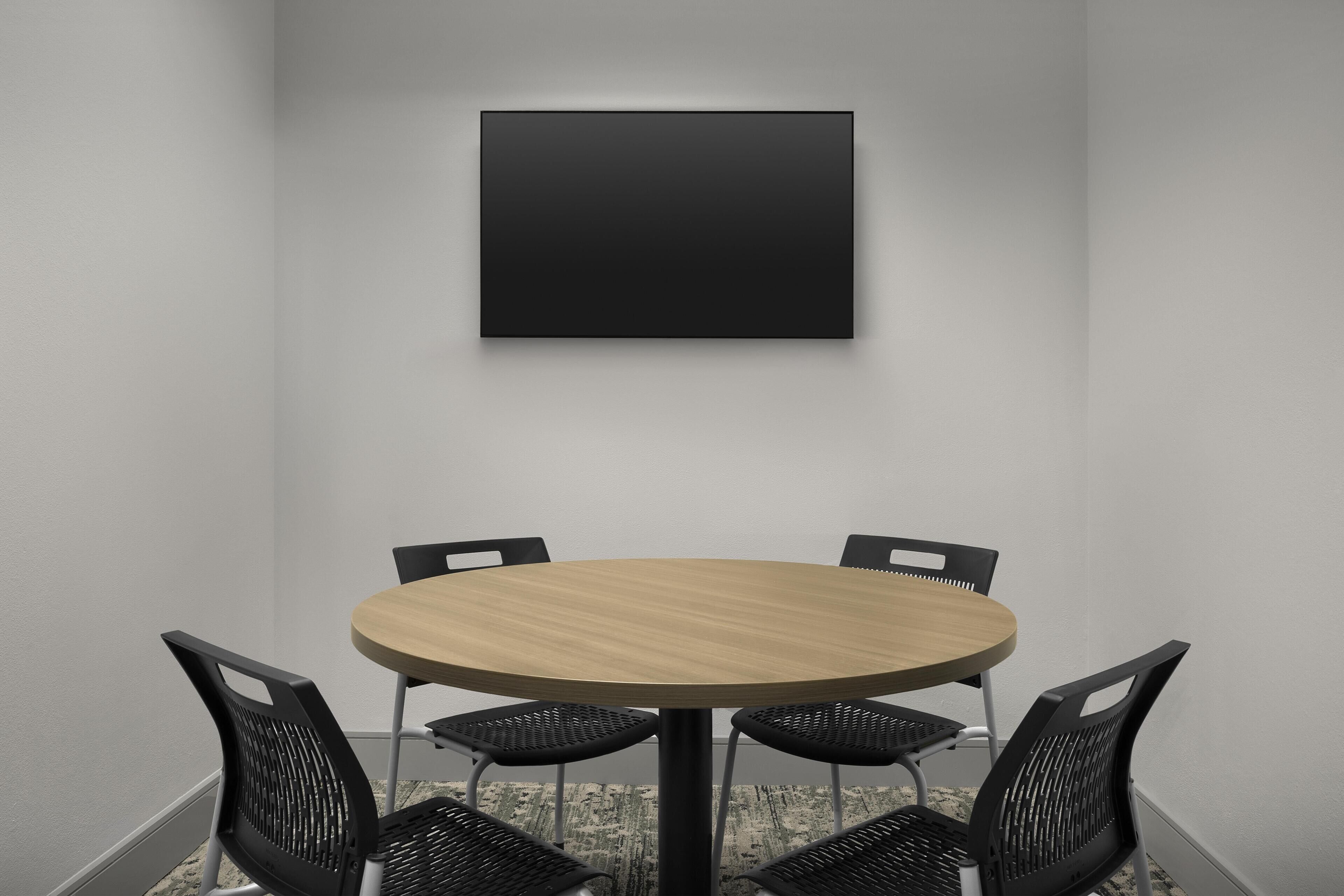 A conference room with a round table, four chairs, and a black framed picture on the wall.