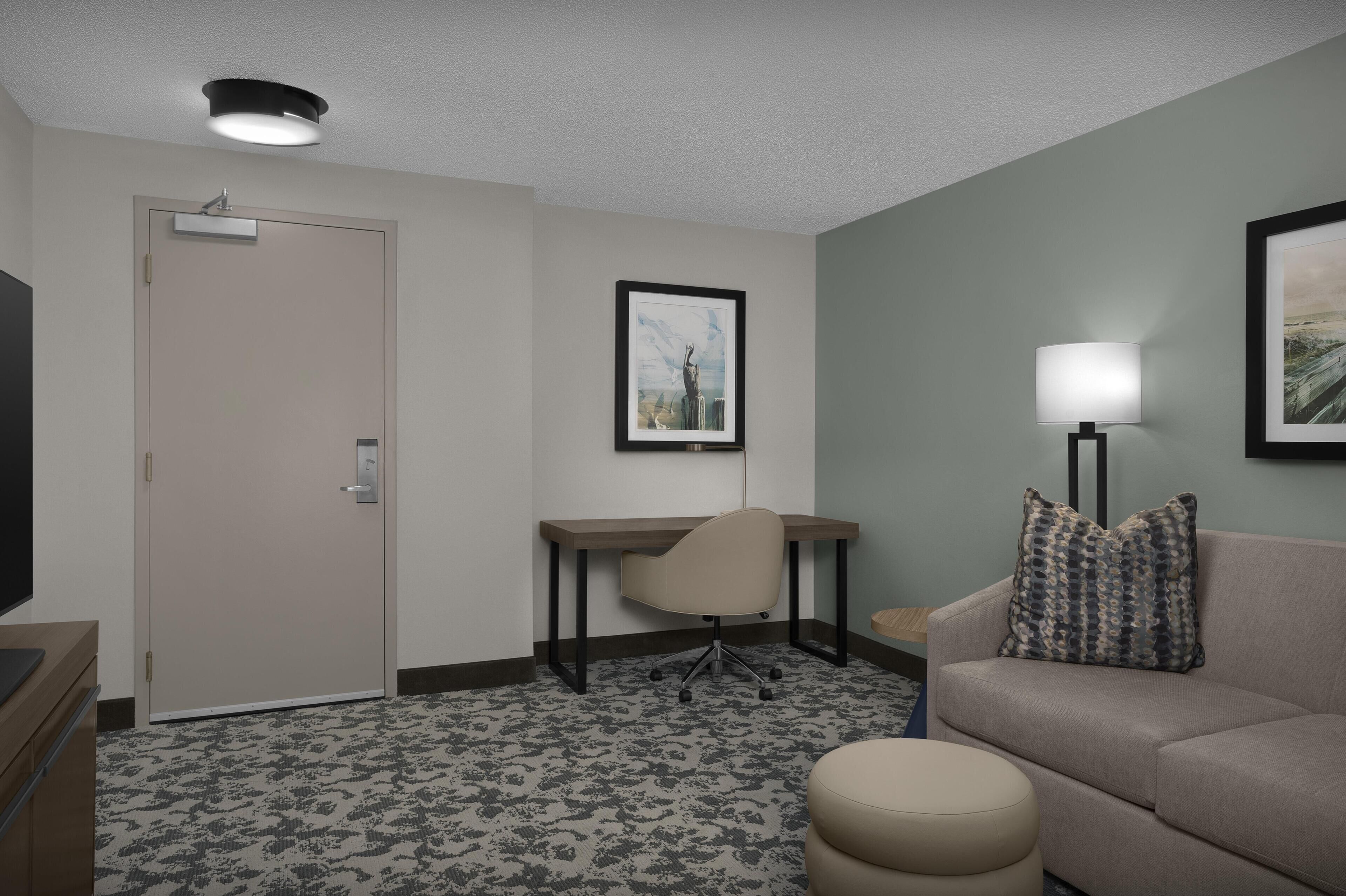 A hotel room with a desk, chair, couch, ottoman, and artwork on the walls.