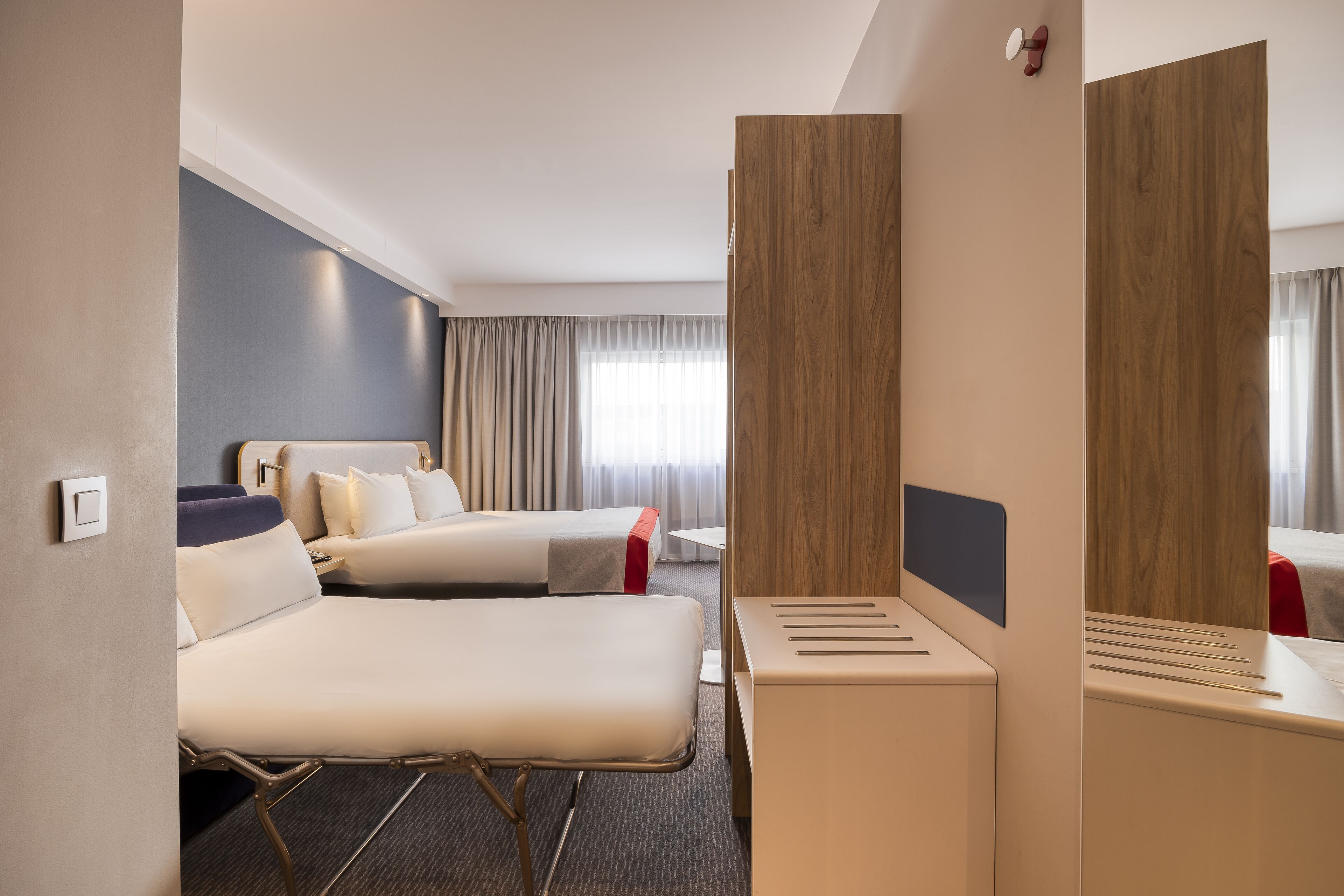 A hotel room with two beds, a small table, a mirror, and a window with curtains.