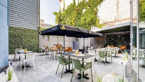 Un patio al aire libre con mesas y sillas, algunas bajo sombrillas, y un muro de ladrillo al fondo.