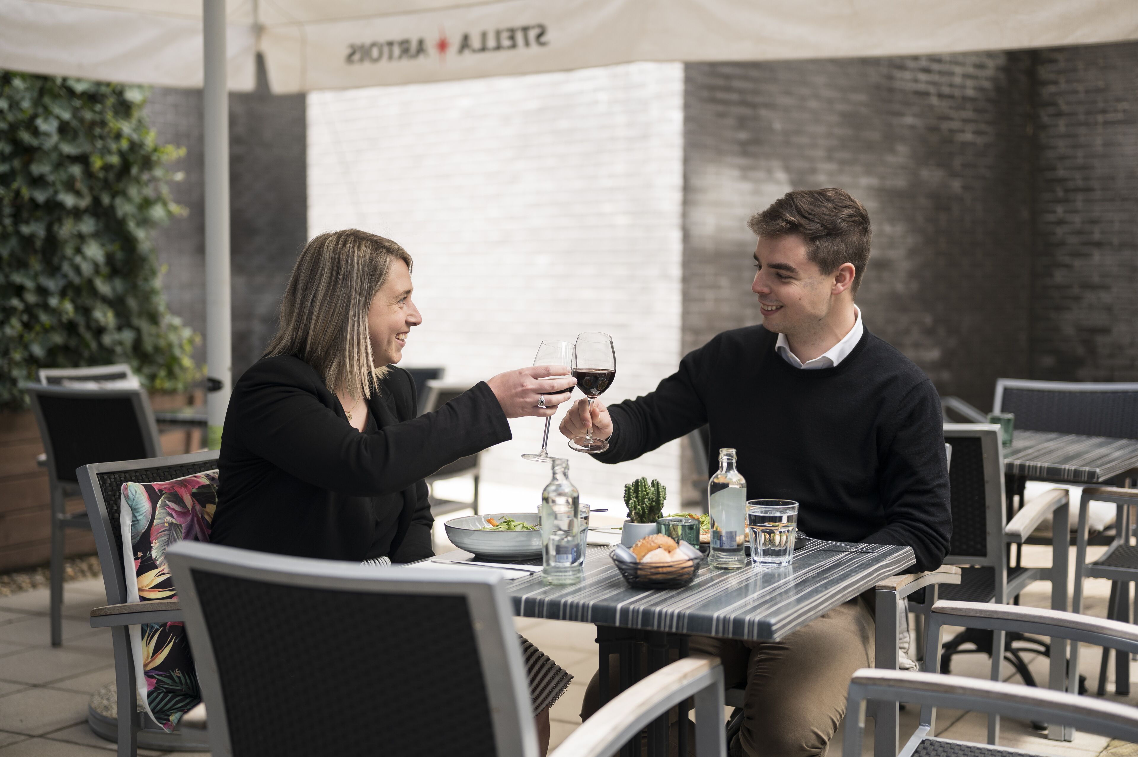Two people toasting with wine glasses at an outdoor café with a brick wall and greenery in the background.