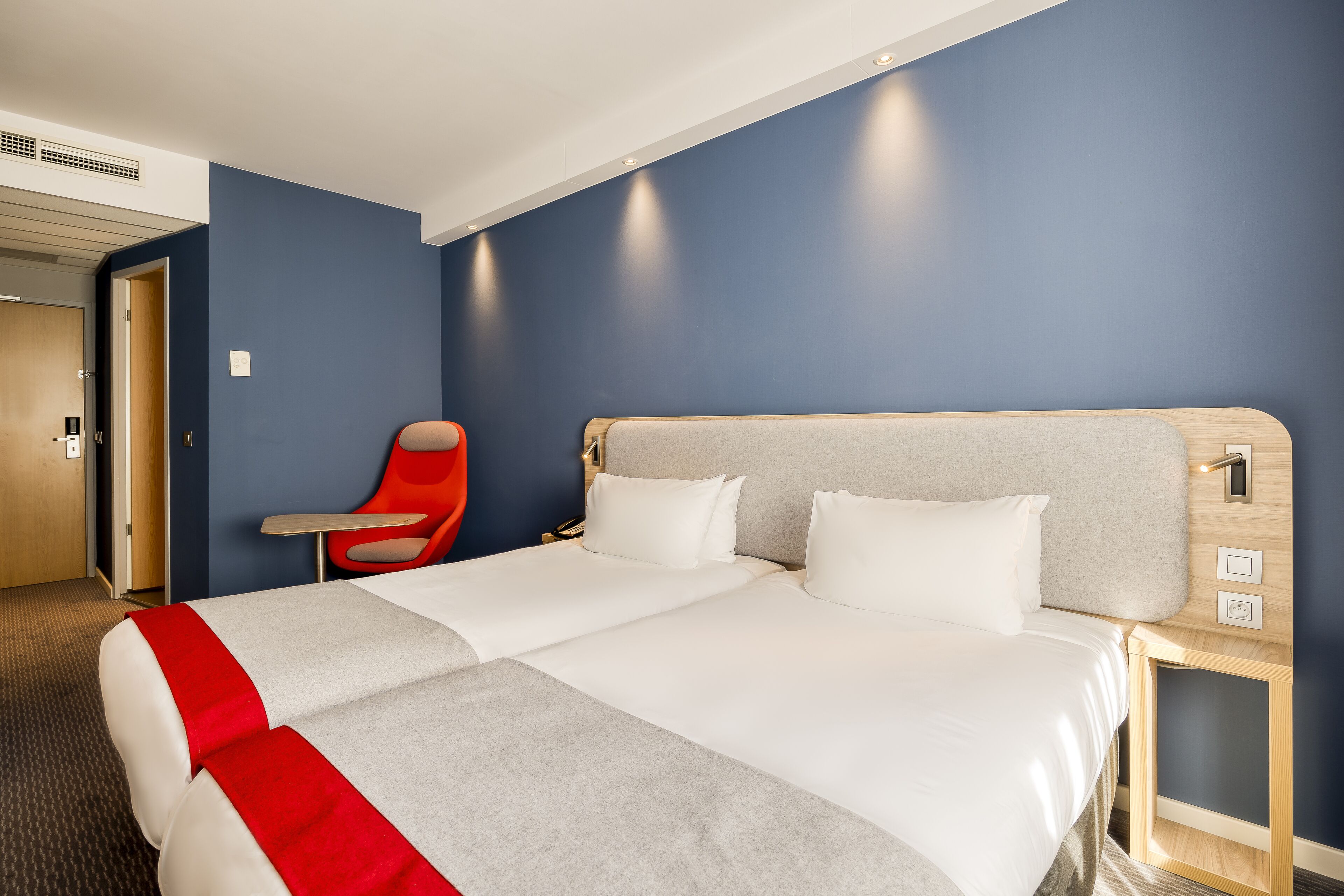 Una habitación de hotel con cama doble, una silla roja, una mesita de noche y un tocador.