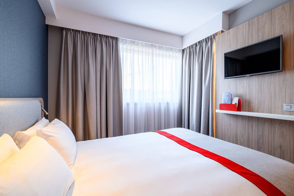 A hotel room with a bed, curtains, a TV, and a shelf with a red object.