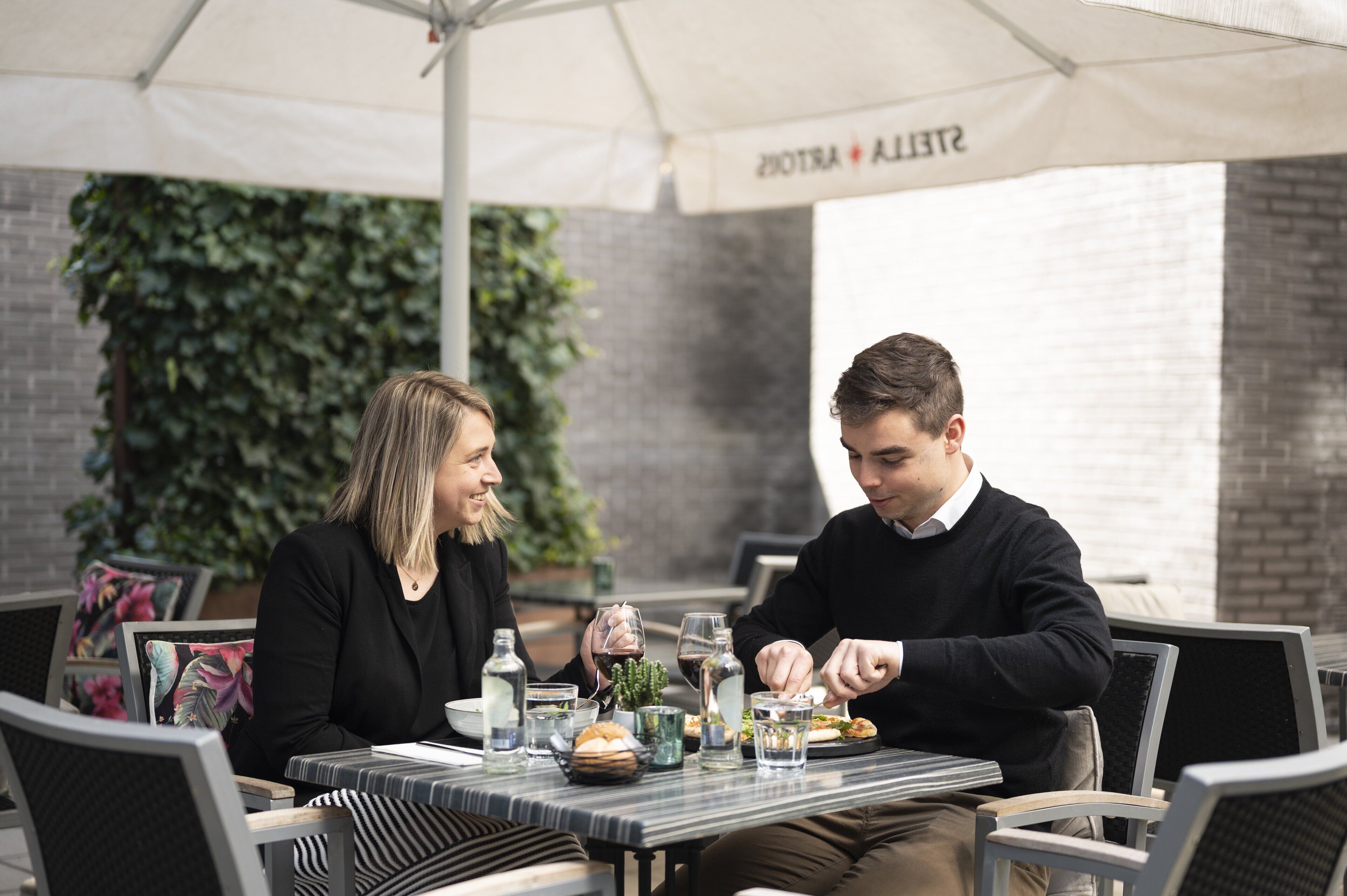 Twee personen aan het dineren aan een buitentafel onder een witte parasol, omringd door stoelen en een bakstenen muur.