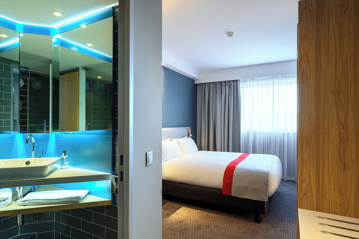 A hotel room with a large bed, a bathroom with a mirror and sink, and a window with curtains.