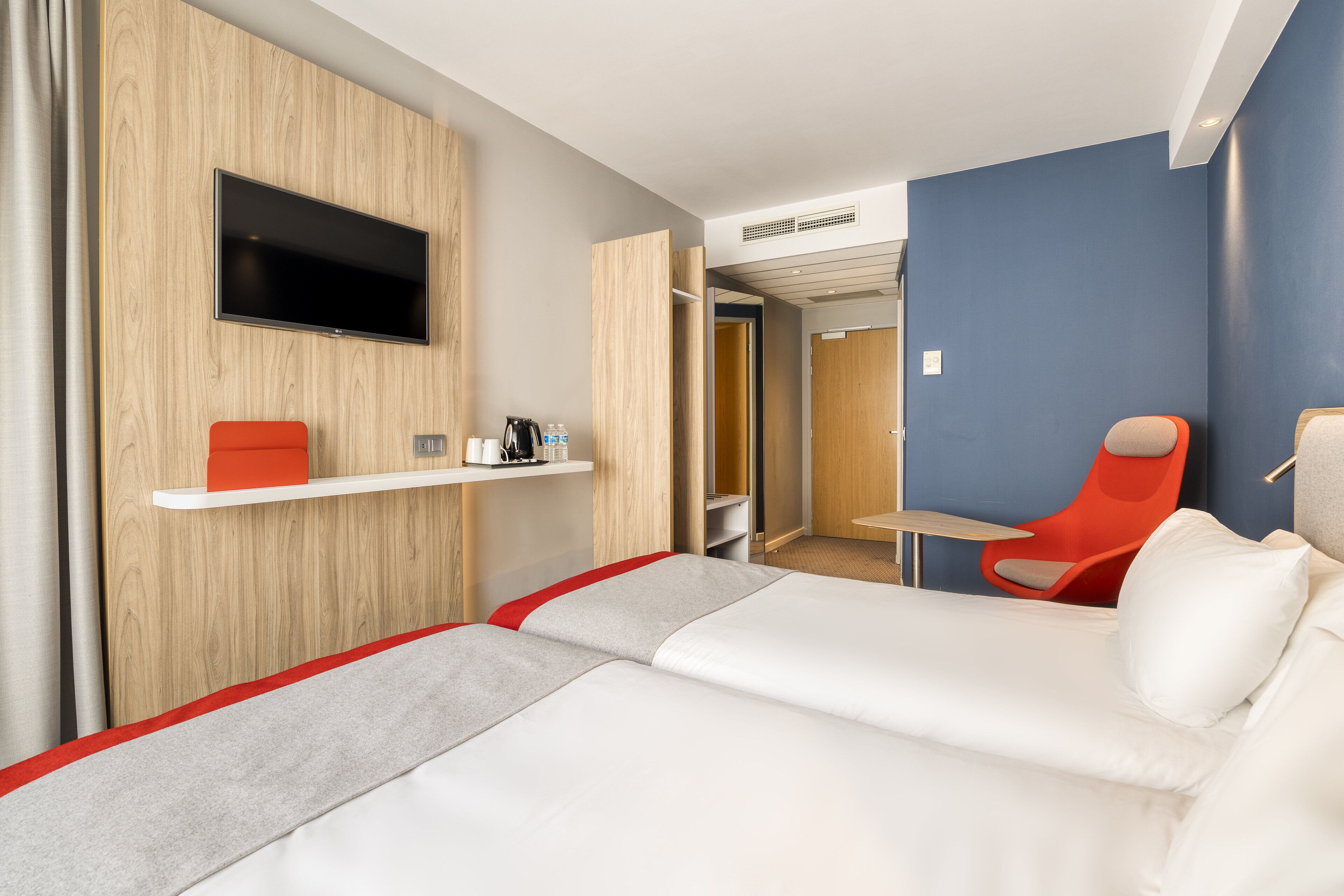 A hotel room with a bed, a TV mounted on the wall, a red chair, and a small table.