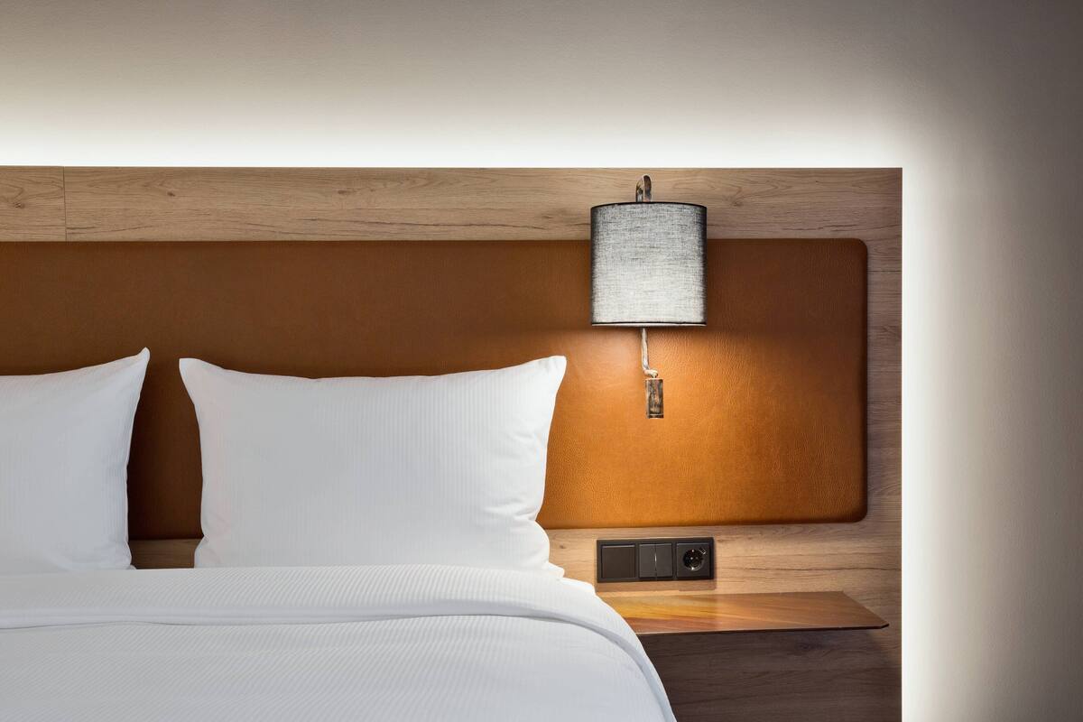 Un lit bien fait, avec du linge de lit blanc, une tête de lit en bois et une lampe fixée au mur.