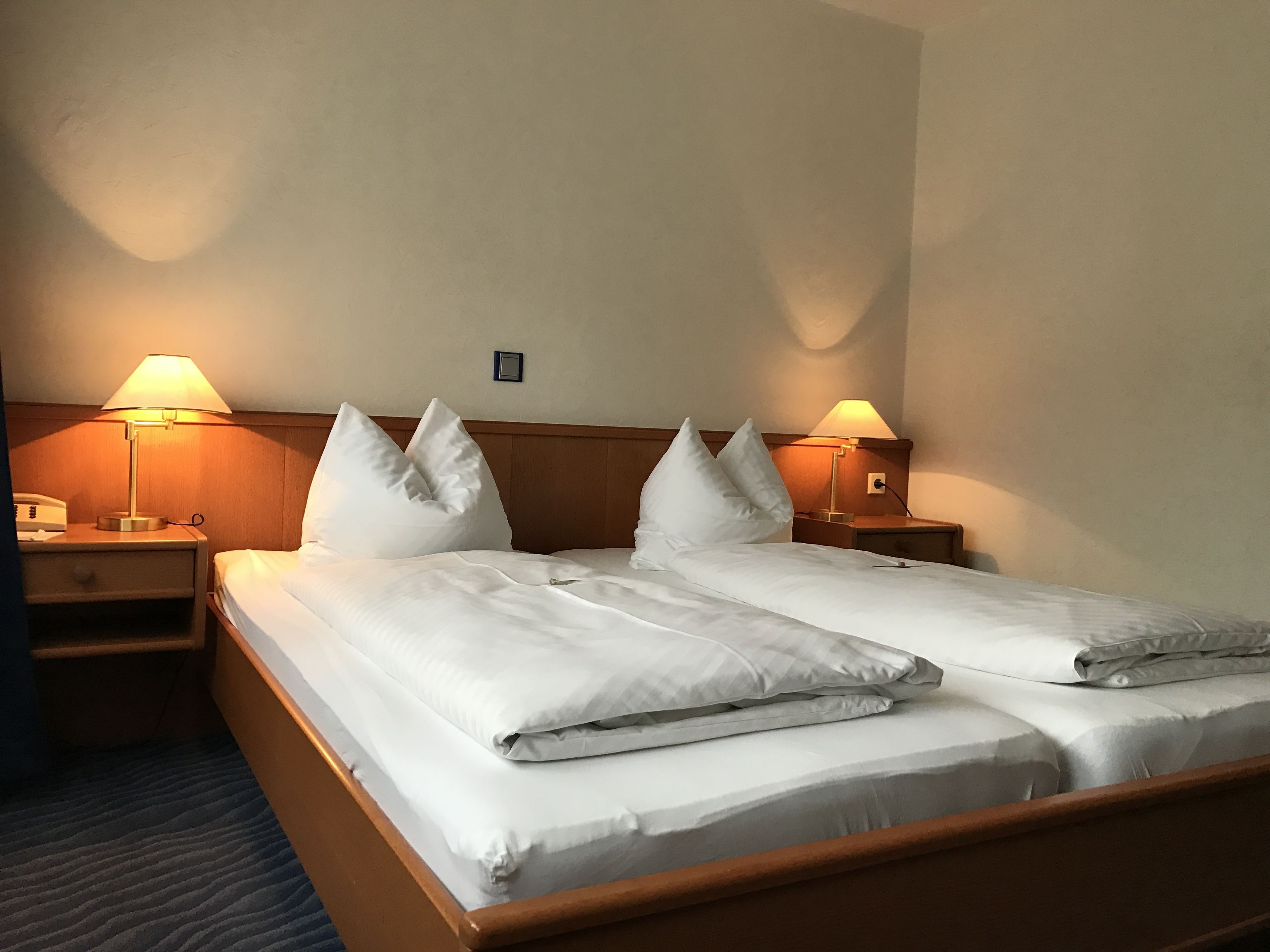 Quarto de hotel com duas camas, cada uma com roupa de cama e almofadas brancas, duas luminárias de cabeceira e um painel de madeira.