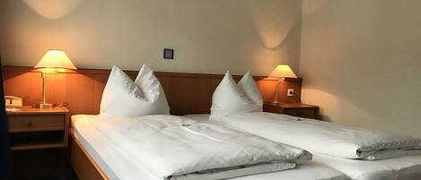 Quarto de hotel com duas camas, cada uma com roupa de cama e almofadas brancas, duas luminárias de cabeceira e um painel de madeira.